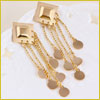 Gold earrings with square tops, hanging chains, and round coin-like charms displayed on a white textured surface.